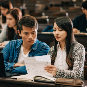 Two students sitting in a classroom, one reading notes, while the other observes intently, surrounded by a group of classmates.